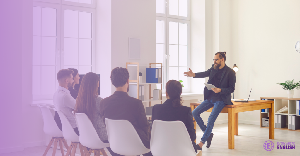 A man sits on a desk, speaking to a group of five people seated in white chairs in a bright, modern office space.