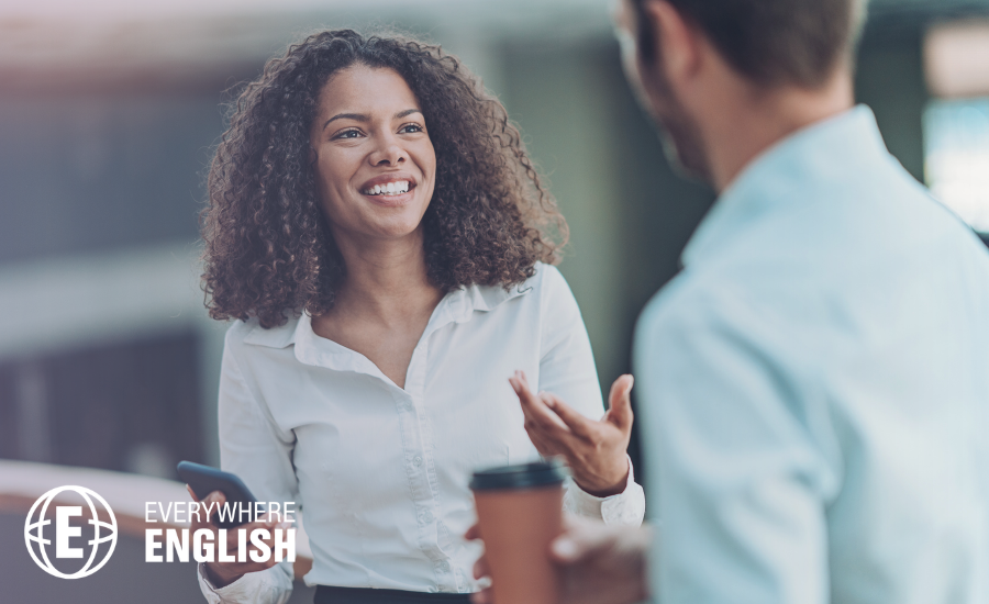 A woman holding a mobile phone smiles and chats with a man holding a coffee cup in a brightly lit indoor setting, discussing The 10 Must-Know Business English Terms for 2026.