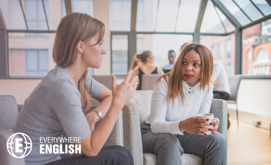 Two women sit on a settee in an office, having a focused conversation. One gestures with her hand while using diplomatic language for difficult conversations at work; the other listens, holding a cup. Other people are talking in the background.