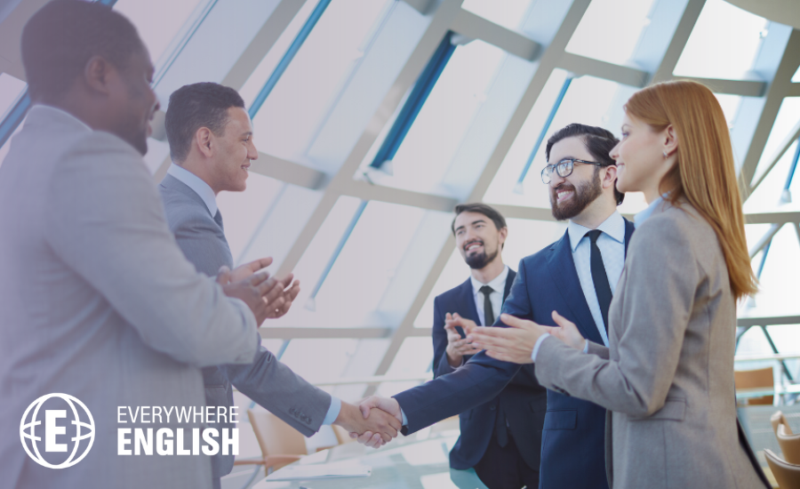 Five business professionals in formal attire stand in an office, two shaking hands whilst others applaud, highlighting the benefits of learning Business English amid large windows in the background.