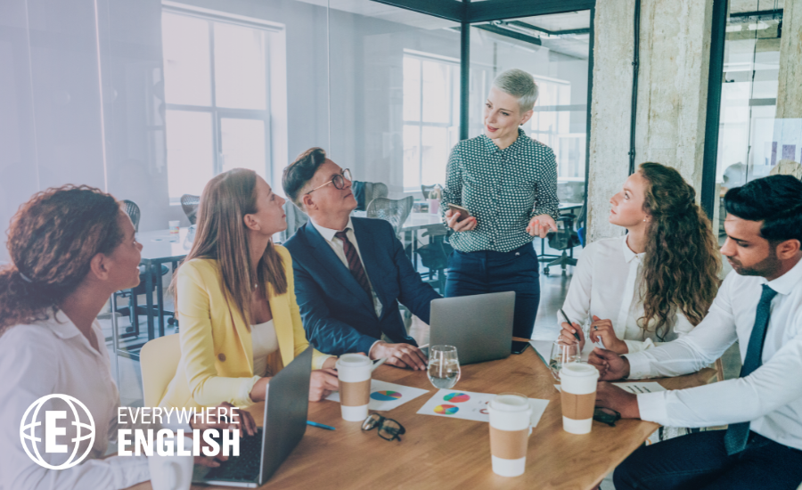 Six people in business attire sit around a conference table with laptops and papers as one person stands to present, sharing tips from "10 Phrases to Sound Natural and Professional in Presentations" in an office setting.