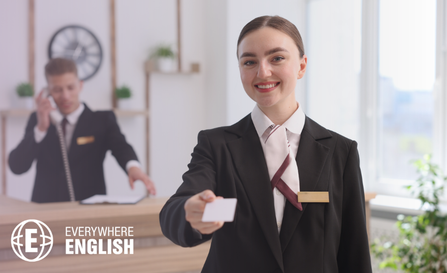 A hotel receptionist in uniform smiles and hands a key card to a guest, whilst another staff member who has completed English for hospitality training talks on the phone in the background.