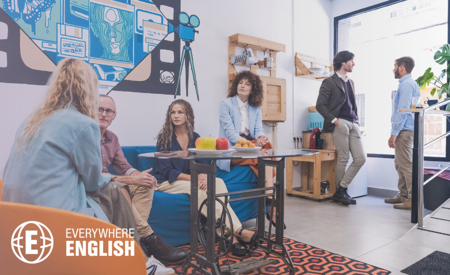 Six people are talking in a casual office lounge with tables, chairs, and wall art; some are seated at a table with apples and snacks, discussing Top 10 English Phrases for Everyday Conversations, whilst others stand near a window.