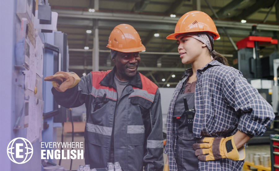 Two factory workers wearing hard hats and protective clothing stand by machinery; one points at a control panel while the other listens attentively, highlighting the benefits of knowing English Language for Manufacturing on the shop floor.